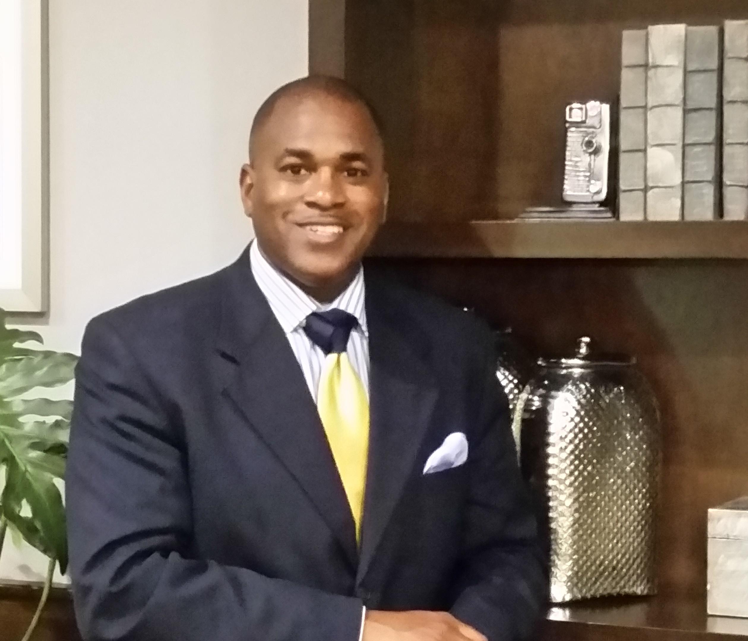 Smiling man in navy suit with yellow tie and white pocket square, seated in front of a wooden bookshelf with decorative it...