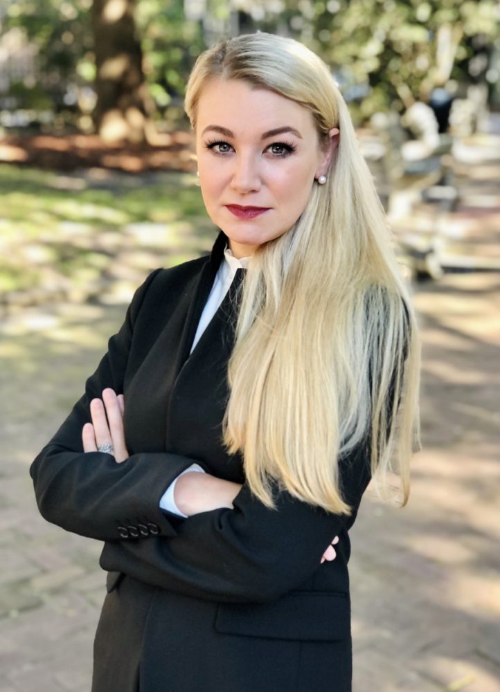 Blonde woman in black blazer and white shirt standing with arms crossed in a tree-lined outdoor setting, professional head...