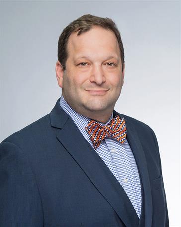 Man in navy blazer and patterned orange and blue bow tie smiling in a professional headshot against a gray background