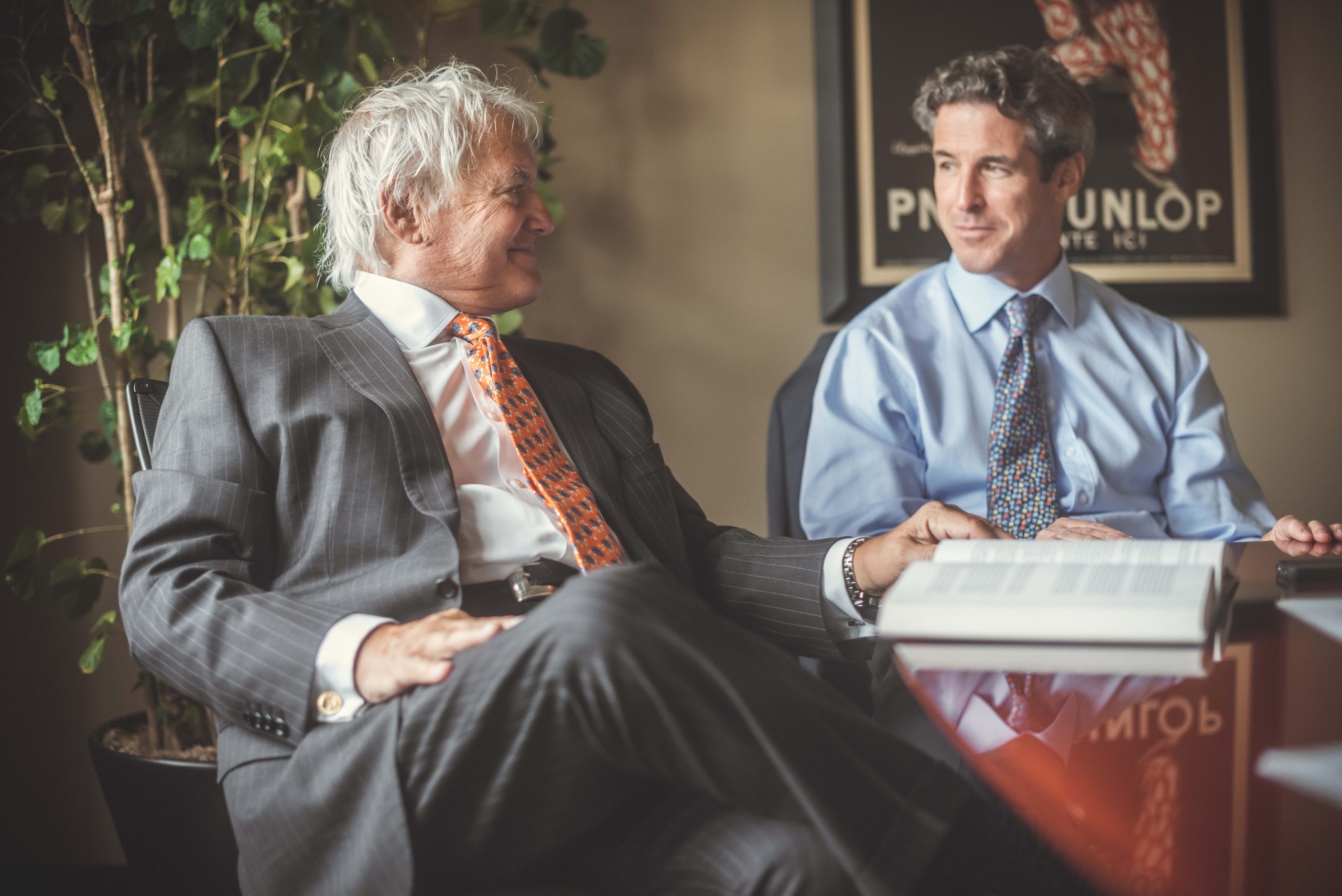 Two suited men in ties having a professional discussion in an office, with an open book on the table and a Dunlop poster o...