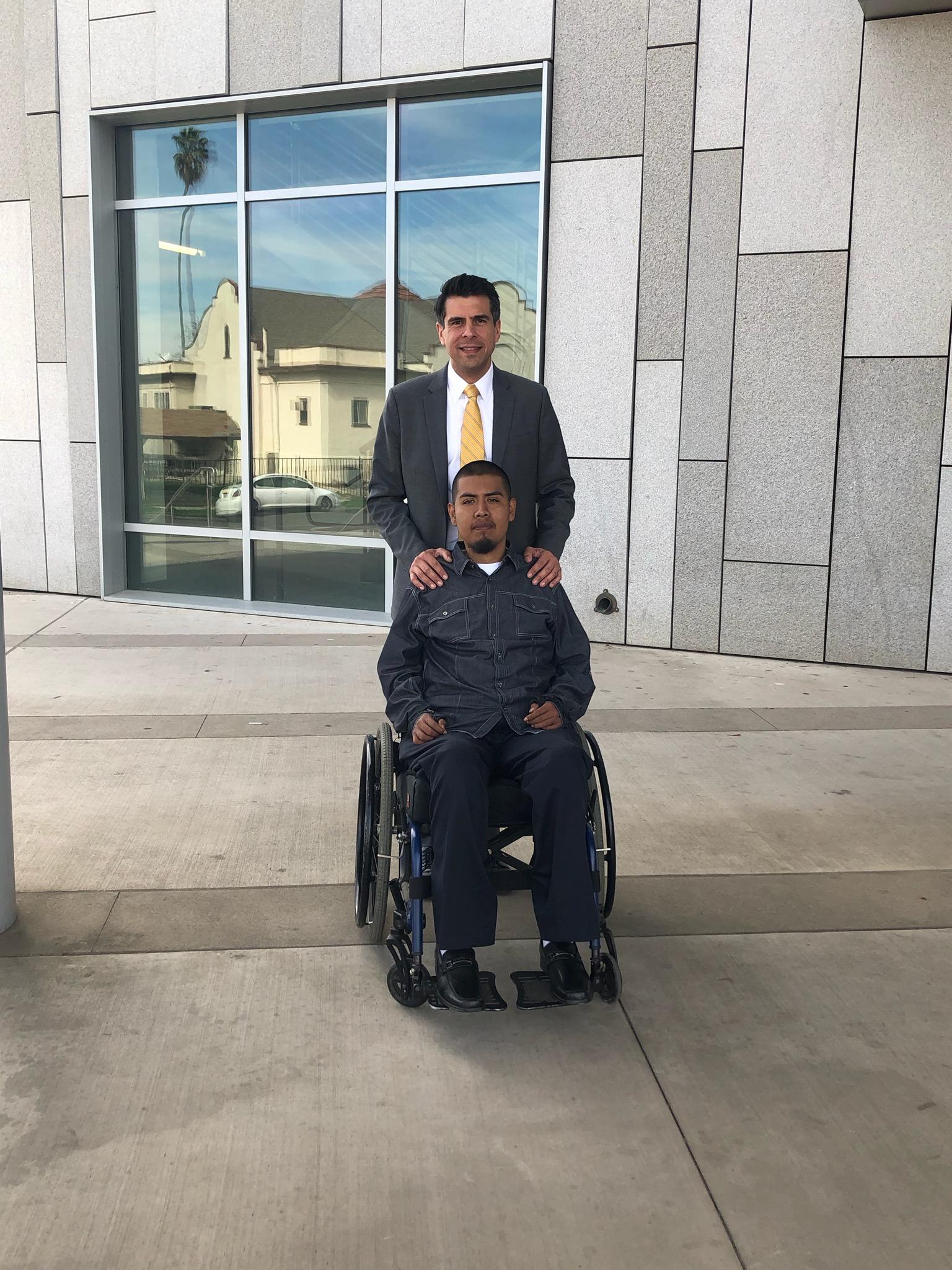 Man in gray suit and gold tie standing behind another man seated in a wheelchair outside a modern stone building.