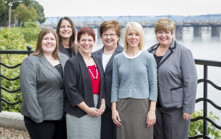 Seven professional women in business attire smiling together outdoors near a river with a bridge in the background.