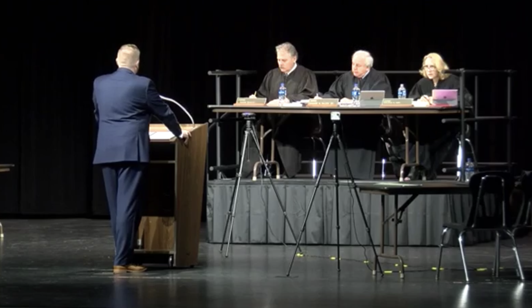 Man in blue suit speaking at podium before three robed judges seated at a draped table on a darkened stage.