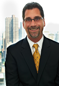 Man in black suit and gold tie smiling, with city skyline visible through window behind him