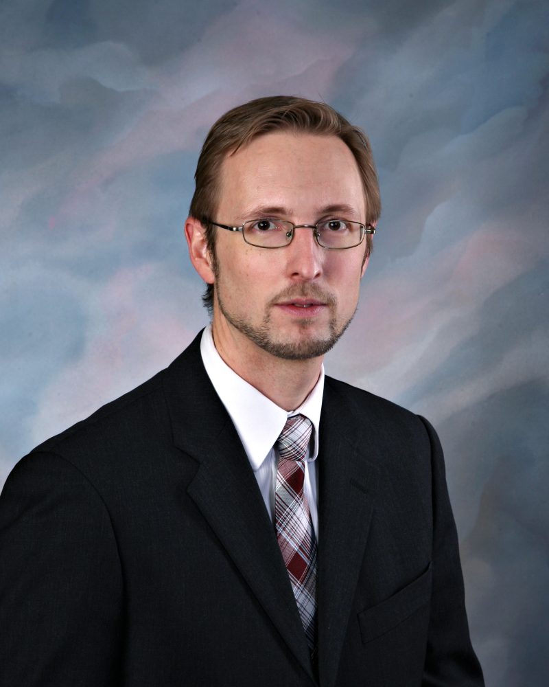 Man with brown hair, glasses, and short beard wearing a black suit and plaid tie against a soft blue-gray studio background.