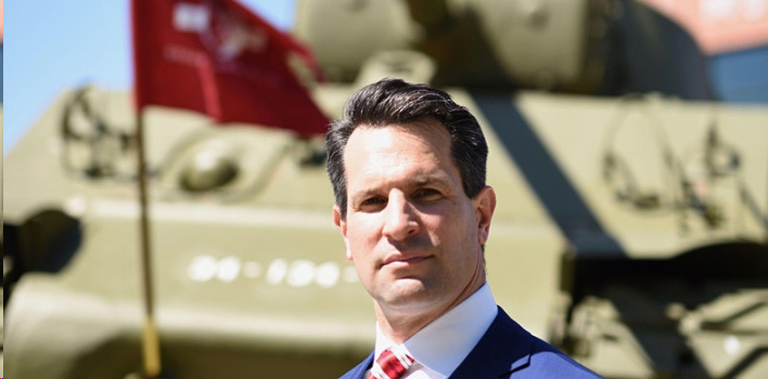 Man in blue suit and red patterned tie standing in front of an olive green military tank with a red flag in the background