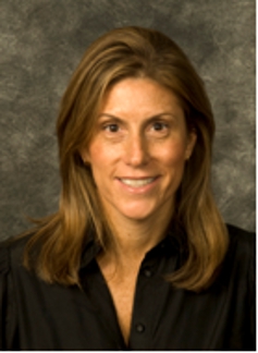 Woman with shoulder-length brown hair wearing a black top, smiling against a gray studio background in a professional head...