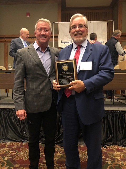 Two men smiling at an awards event, one holding a plaque reading "J. Kevin Holmes" near an "Injured Workers" banner.