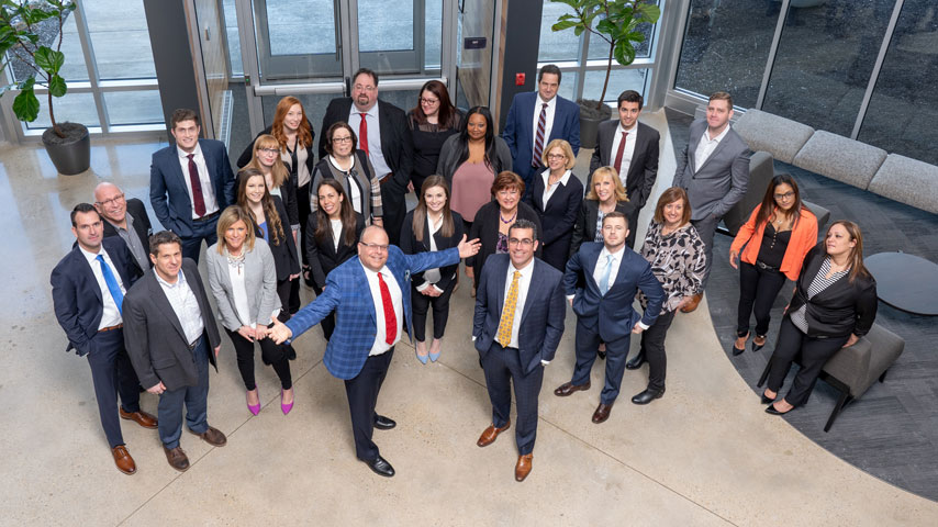 Large group of approximately 25 business professionals in suits posing together in a modern office building lobby, aerial ...