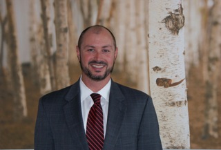 Smiling man with short beard wearing a gray suit and red striped tie, standing in front of a birch tree backdrop.