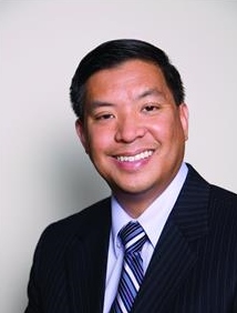 OC Wills and Trusts Responds to Covid-19 Virus 1 Smiling Asian man in a dark pinstripe suit and striped tie against a light gray background, professional headshot.