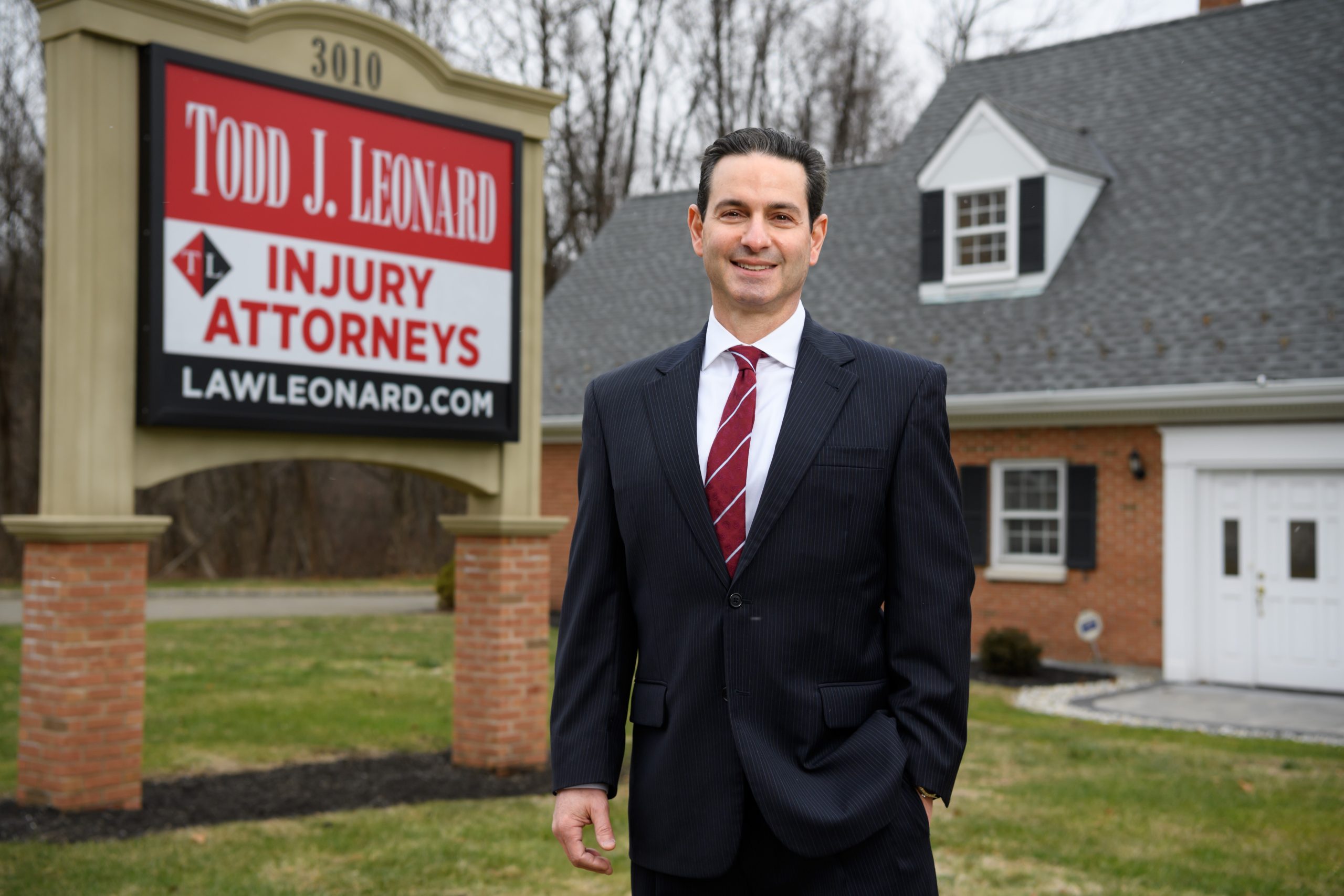 Man in dark suit and red striped tie standing before Todd J. Leonard Injury Attorneys sign at 3010, lawleonard.com, brick ...