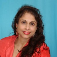 Smiling South Asian woman with long dark hair wearing a red top and red earrings against a teal background