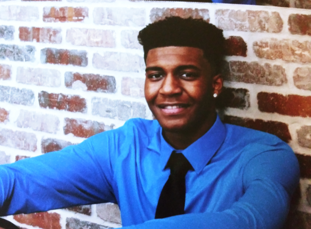 Herman Law, P.A. Awards Inaugural Second Chance Scholarship 3 Young man smiling in a blue dress shirt and black tie, leaning against a rustic brick wall in a formal portrait photo.
