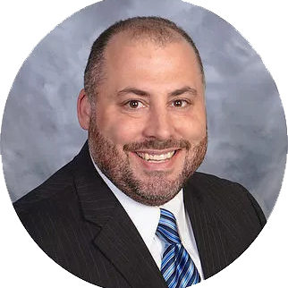Smiling man with short dark hair and beard wearing a black suit jacket, white shirt, and blue striped tie against a gray b...