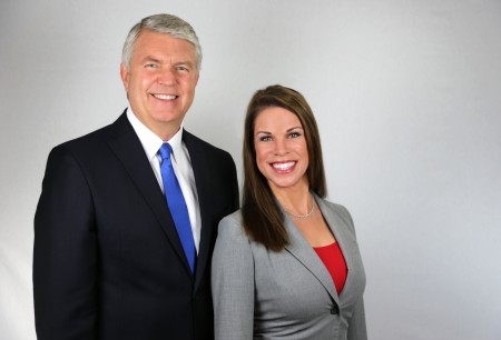 Man in black suit with blue tie and woman in gray blazer with red top smiling together in a professional portrait.