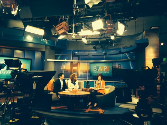 Three people seated around a table on a TV studio set with BAP Bay Area People signage and studio lights overhead.