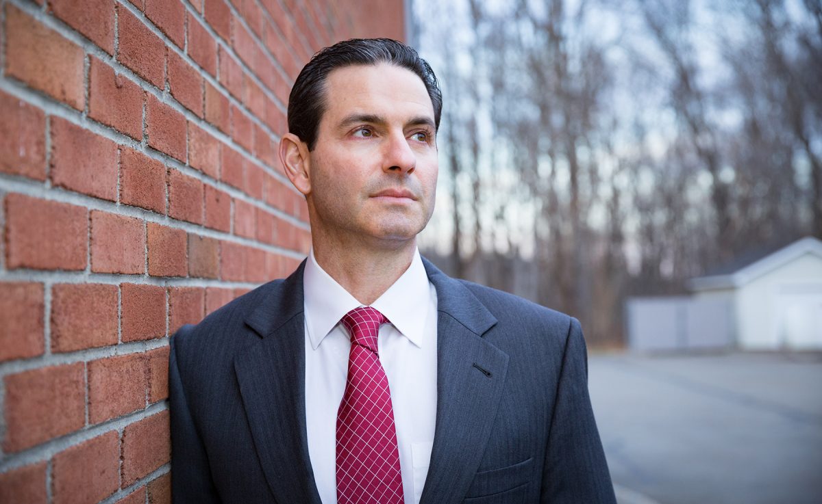 Man in dark gray suit and red patterned tie leaning against a brick wall, gazing into the distance outdoors.