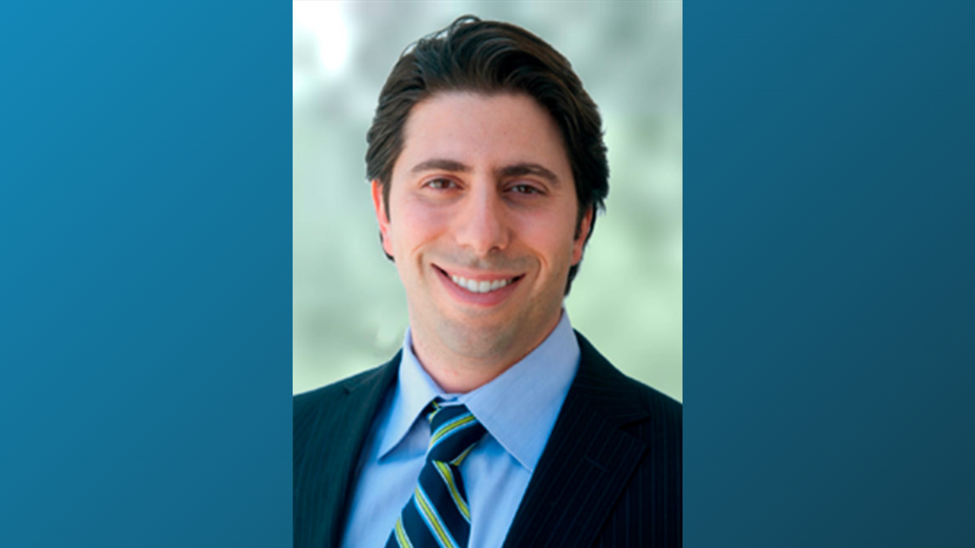 Smiling man with dark hair wearing a navy pinstripe suit, light blue dress shirt, and striped tie against a blurred outdoo...