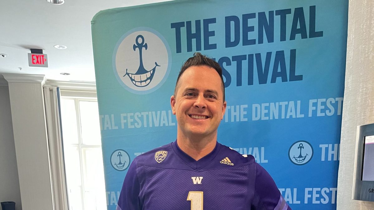 Smiling man wearing a purple Washington Huskies football jersey standing in front of a teal "The Dental Festival" banner w...