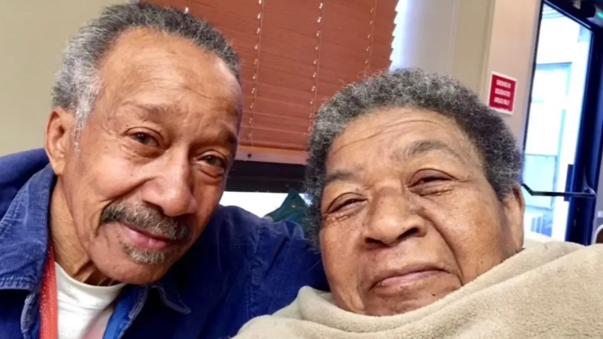Two elderly Black adults posing close together for a selfie, man in blue jacket smiling, woman wrapped in a beige blanket ...