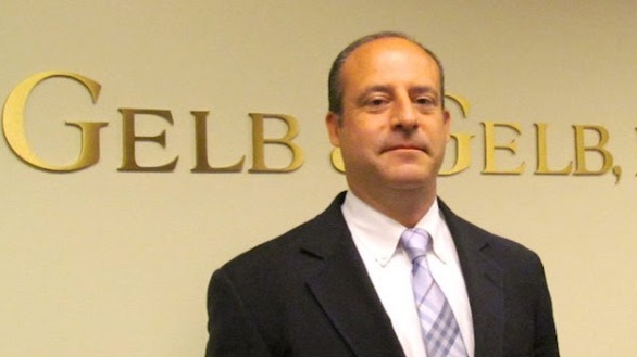 Gelb & Gelb Awards Scholarship for Safe Driving for Fourth Year 11 Man in dark suit and lavender tie standing before gold "Gelb & Gelb" lettering on a beige wall