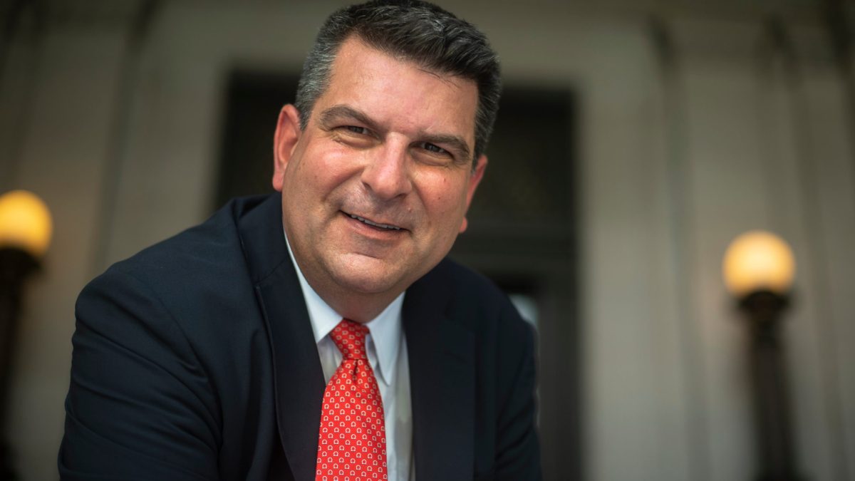 Middle-aged man in dark suit and red tie smiling at camera in a formal indoor setting with blurred columns and wall sconces.