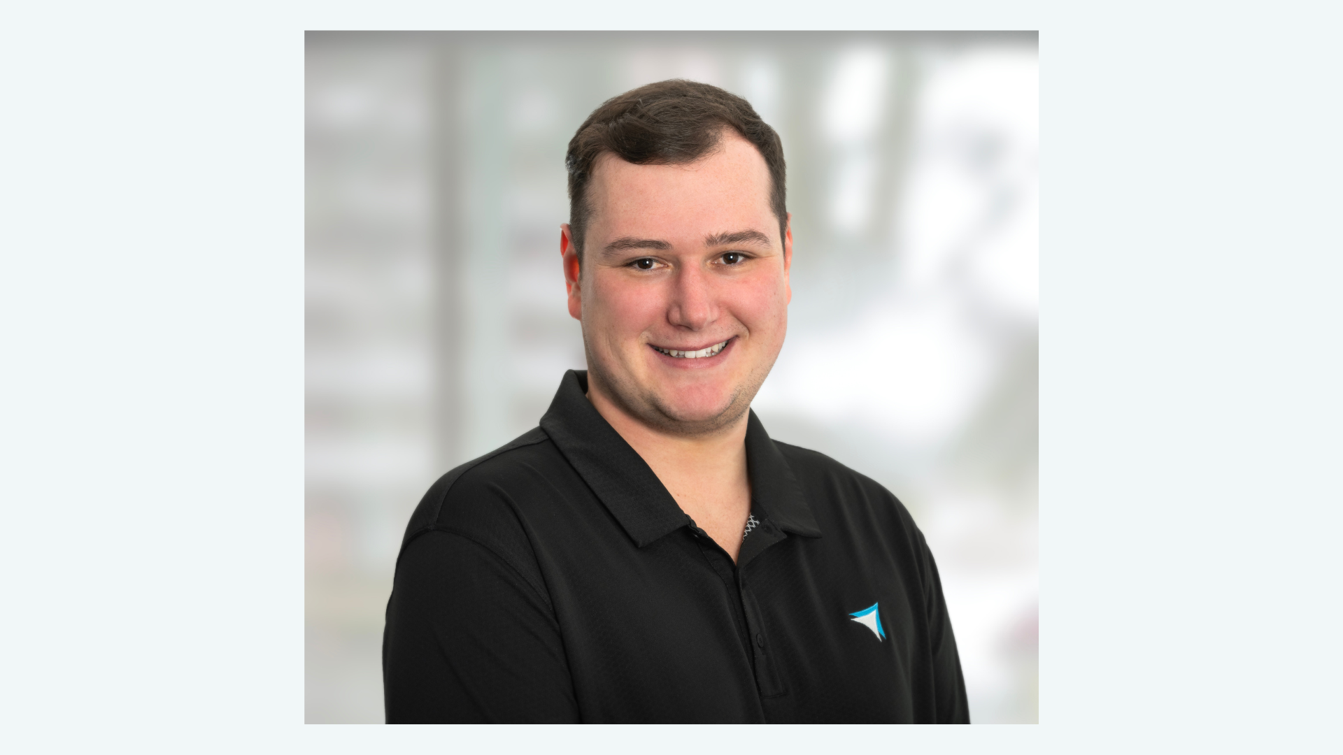 Smiling young man with short brown hair wearing a black polo shirt with a blue and white logo, professional headshot with ...