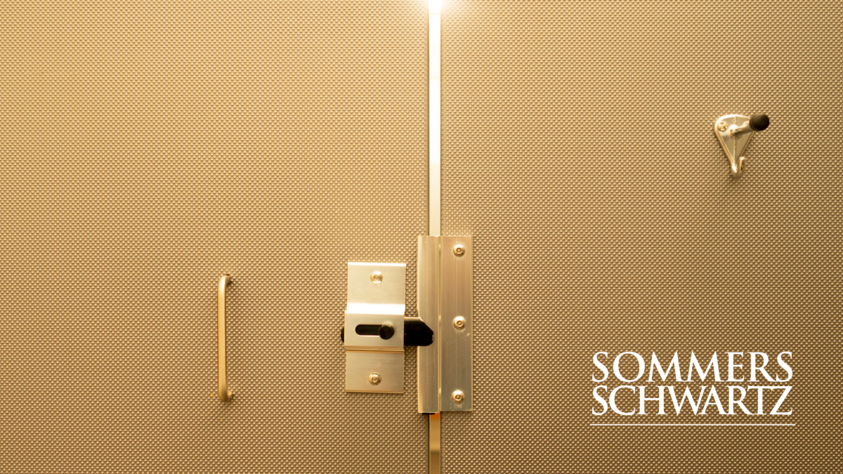 Beige public restroom stall door with gold slide bolt lock and coat hook, Sommers Schwartz logo in bottom right corner