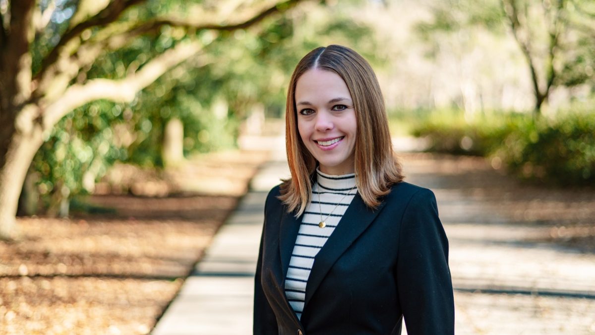 Young woman smiling in a tree-lined park path, wearing a black blazer over a striped turtleneck with a gold necklace.