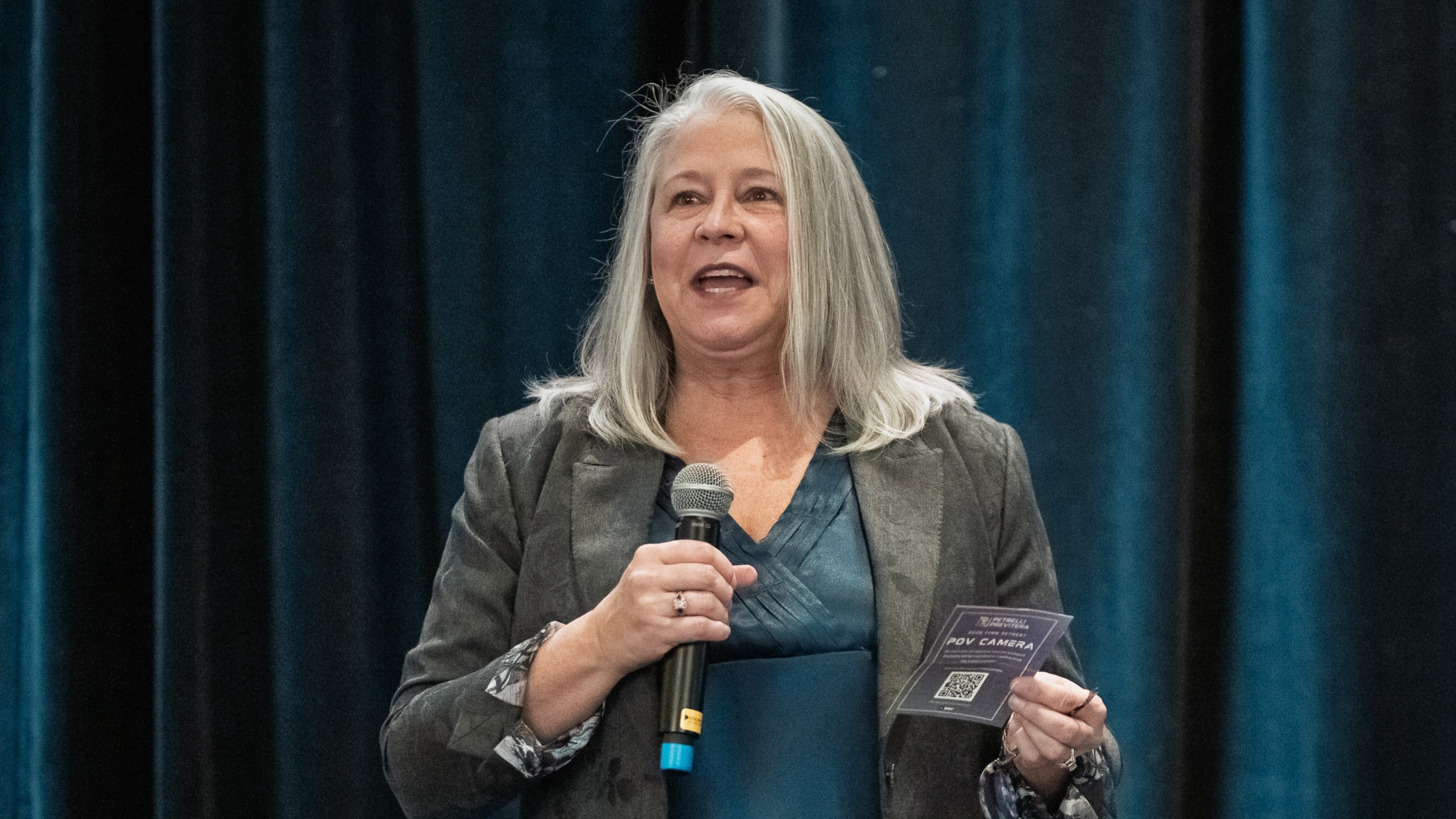 Woman with gray hair speaking into a handheld microphone on stage, holding a small card labeled "POV CAMERA" before a teal...